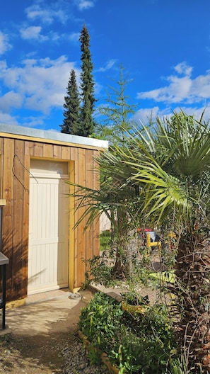 Eine hölzerne Gartenhütte mit einer weißen Tür, umgeben von verschiedenen Pflanzen und einem strahlend blauen Himmel.