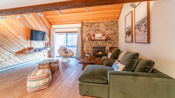 A cozy living room with a stone fireplace, wooden beams, a green sofa, and a flat-screen TV. - Cozy 2+Loft in Snowcreek Meadow w/ Mountain Views! (Mammoth Lakes)