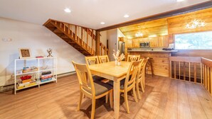 A wooden dining area with a table and chairs, a white shelving unit with boxes, and a staircase leading to a kitchen area with wooden cabinets and a hanging light fixture. - Cozy 2+Loft in Snowcreek Meadow w/ Mountain Views! (Mammoth Lakes)