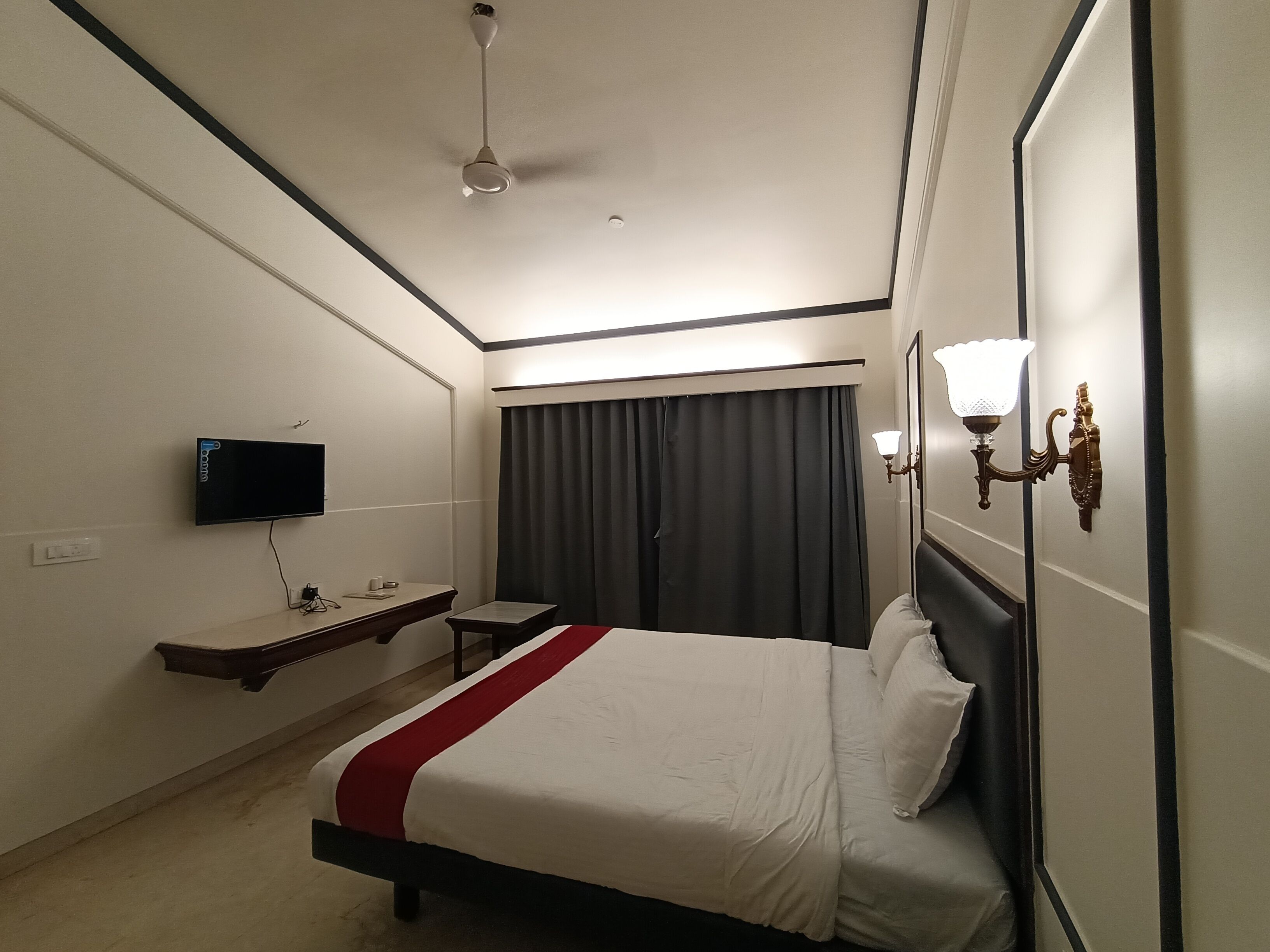 Una habitación de hotel con cama, televisión montada en la pared, escritorio y ventana con cortinas.