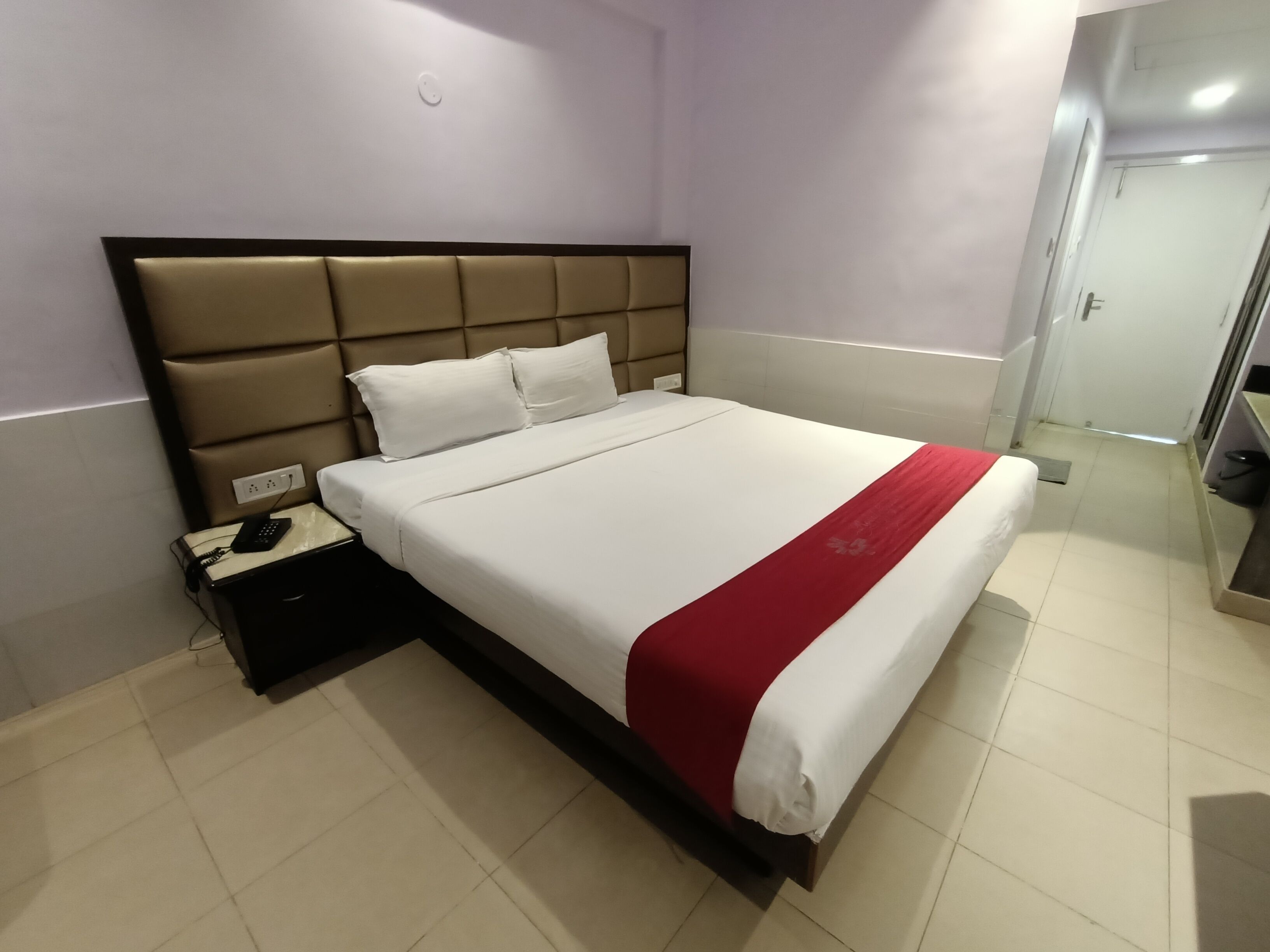 A hotel room with a large bed, a bedside table with a phone and charger, and a red throw blanket.