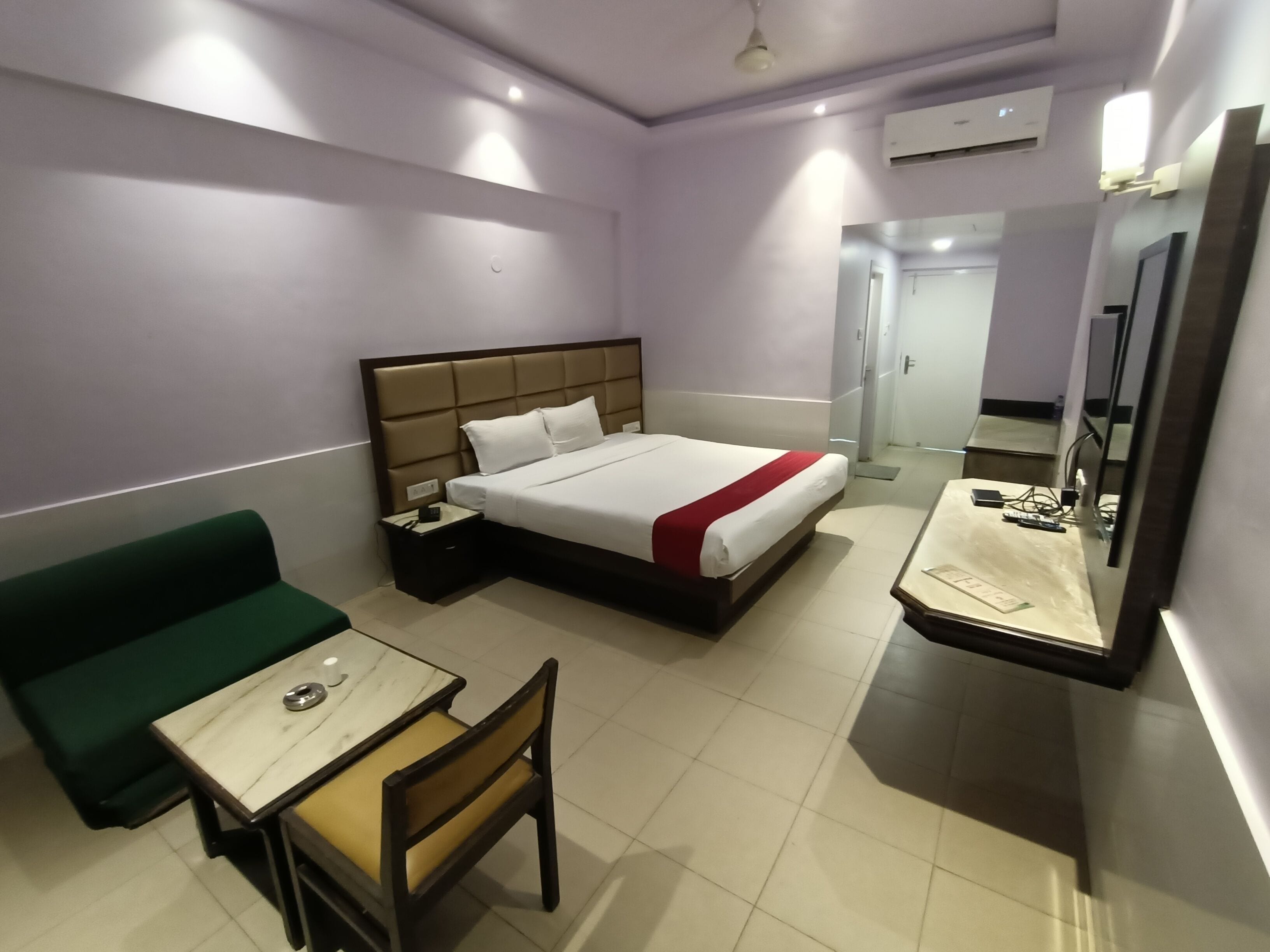 A hotel room with a bed, a desk, a chair, a green sofa, a mirror, and an air conditioning unit.