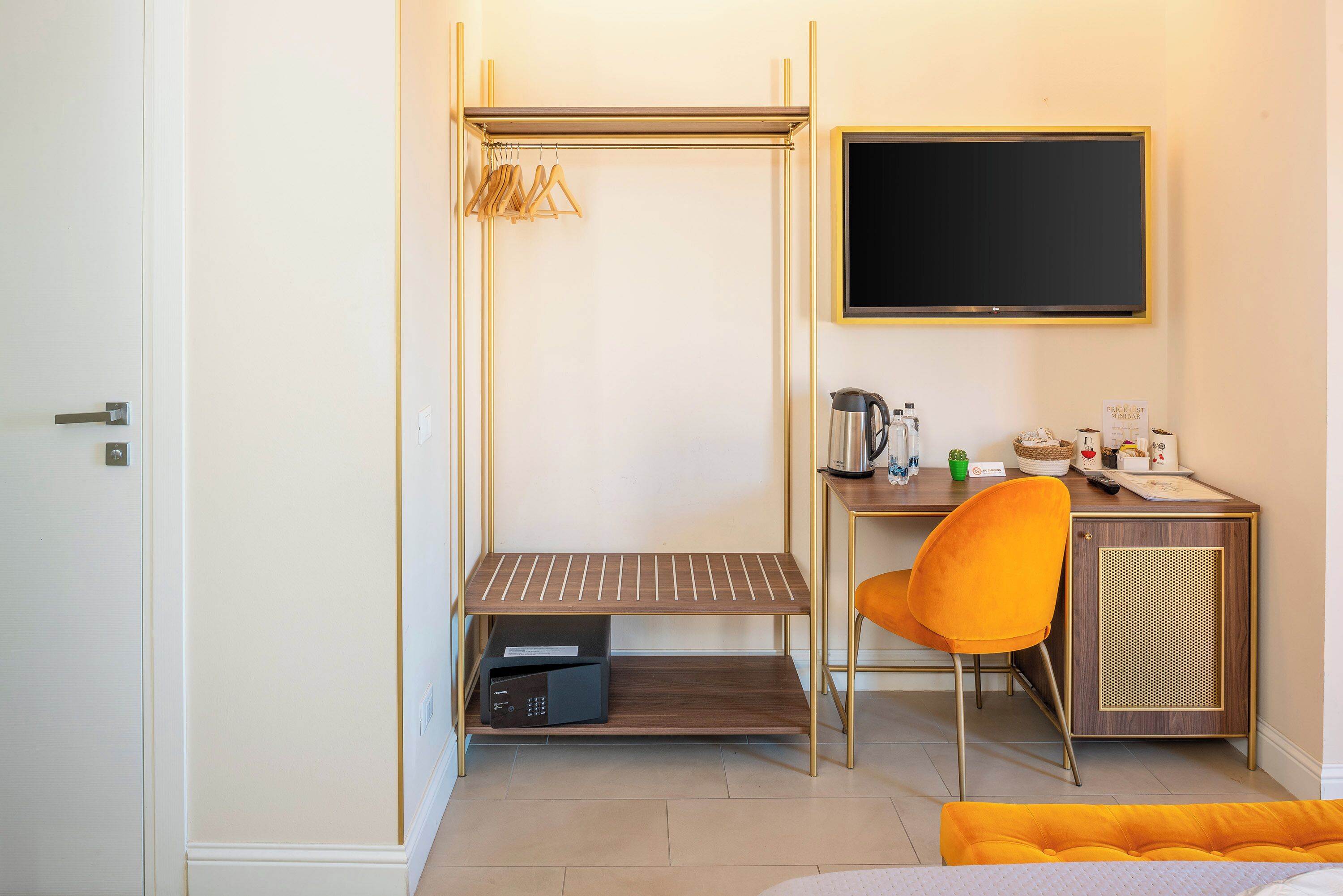 A compact hotel room with a bed, a desk with a chair, a small shelf, a coat rack, and a television.