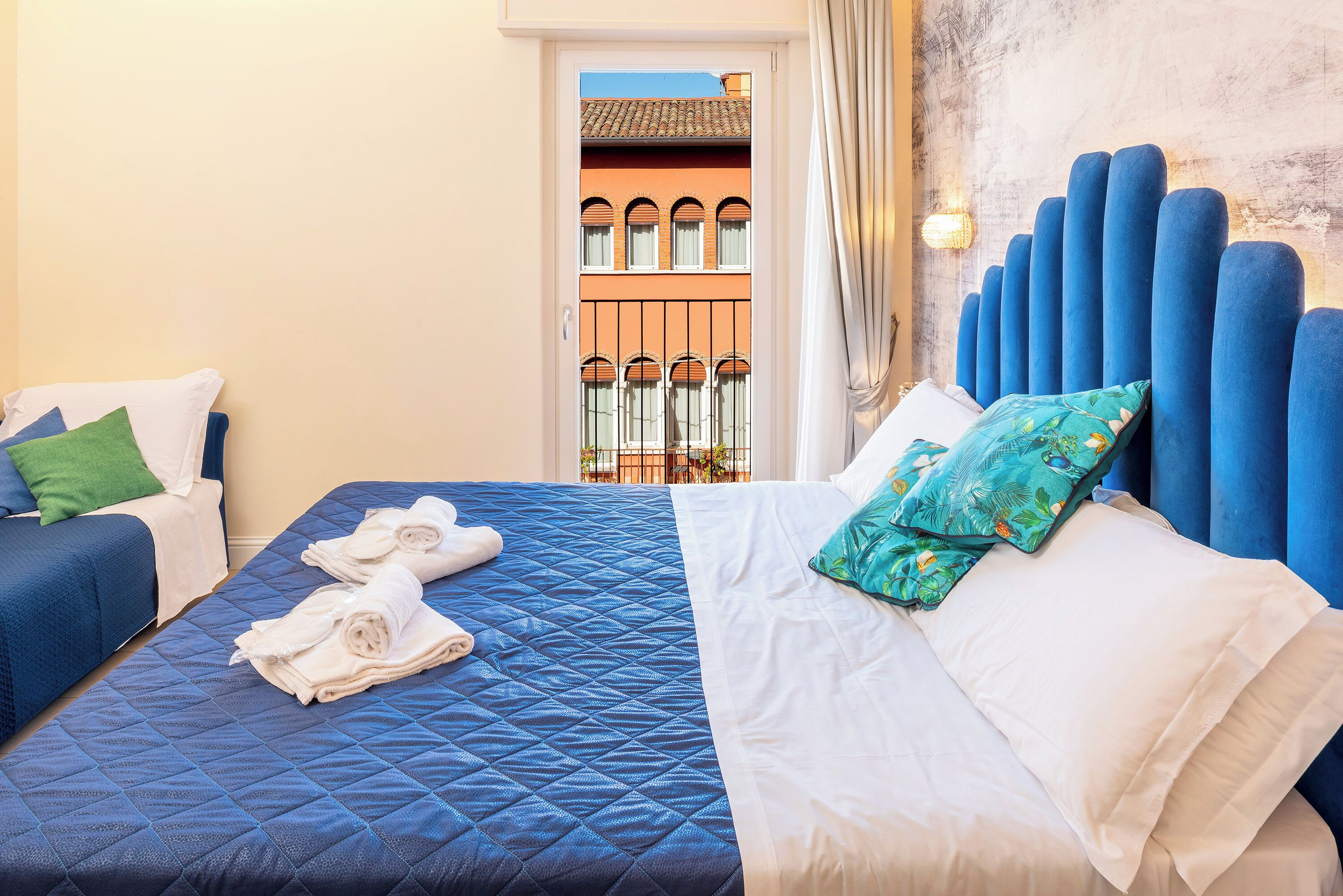 Une chambre d’hôtel avec deux lits, l’un recouvert d’une couette bleue et l’autre de draps blancs. Des serviettes sont posées sur les lits, et on aperçoit, à travers une porte ouverte, un bâtiment doté de fenêtres en arc de cercle.