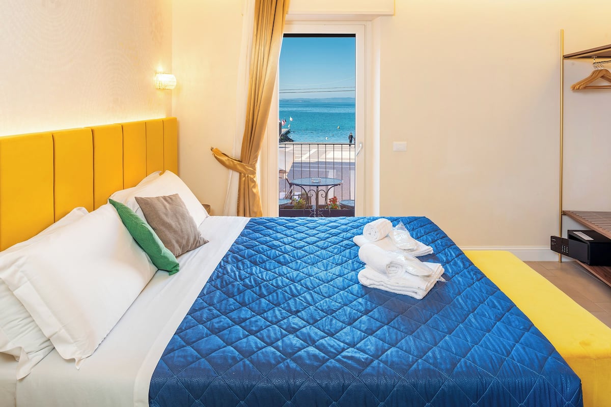 A hotel room with a bed, a balcony with a view of the sea, and a yellow wall.