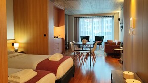 A hotel room with two beds, a dining table with chairs, a sofa, and a view of the city through large windows. - Fervença Apartments - Corgo (Vila Nova de Gaia)