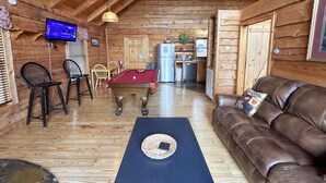 Interior acolhedor de uma cabana com mesa de bilhar, sofá de couro e televisão na parede.