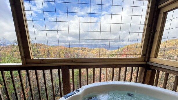 Ein Whirlpool auf einer Holzterrasse mit Blick auf Herbstlaub und Berge durch ein Fenster.