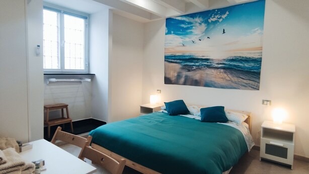 A bedroom with a bed, a desk, and a chair. There is a window and a painting on the wall.