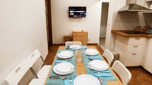 A dining area with a long table set for four, white chairs, a television mounted on the wall, and a kitchenette to the right.