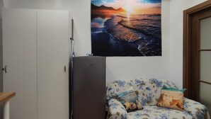 A compact living space with a kitchenette, a sofa with a patterned cover, a wall-mounted artwork depicting a sunset over water, and a door with a glass panel.