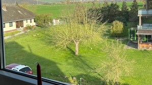 Une vue depuis une fenêtre donnant sur un paysage verdoyant, avec un arbre, un bâtiment et des collines au loin.