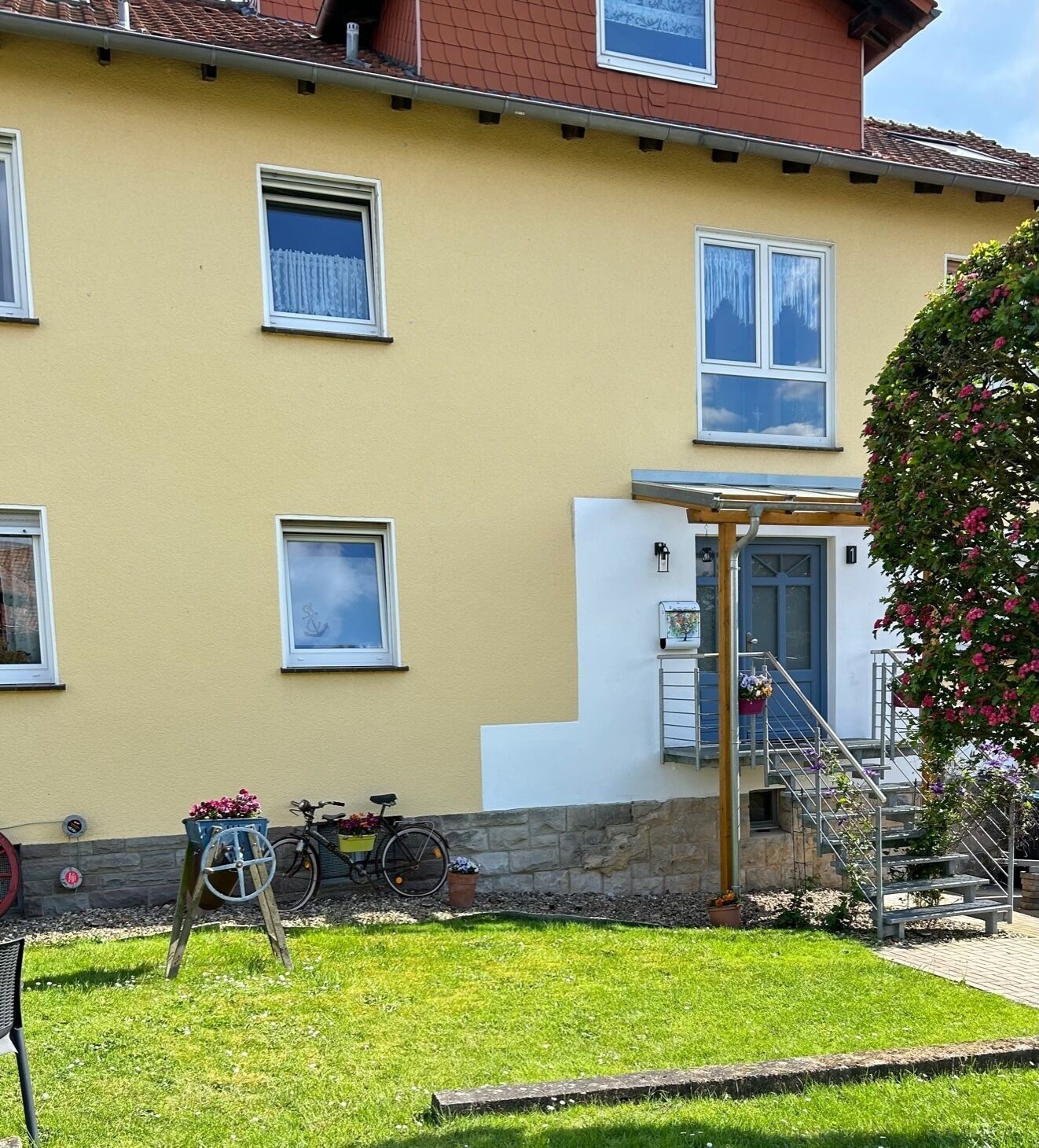Ein gelbes Gebäude mit einer Treppe, die zu einem überdachten Eingang führt, einem draußen abgestellten Fahrrad und einem kleinen Garten.