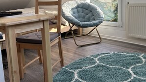 Une pièce avec une table en bois, une chaise rembourrée, un tapis bleu à motifs et une fenêtre donnant sur des arbres.