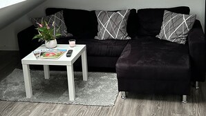 Un salon moderne avec un canapé noir, des coussins blancs et à motifs, une table basse blanche agrémentée de magazines et d’une plante, et une lucarne au plafond.