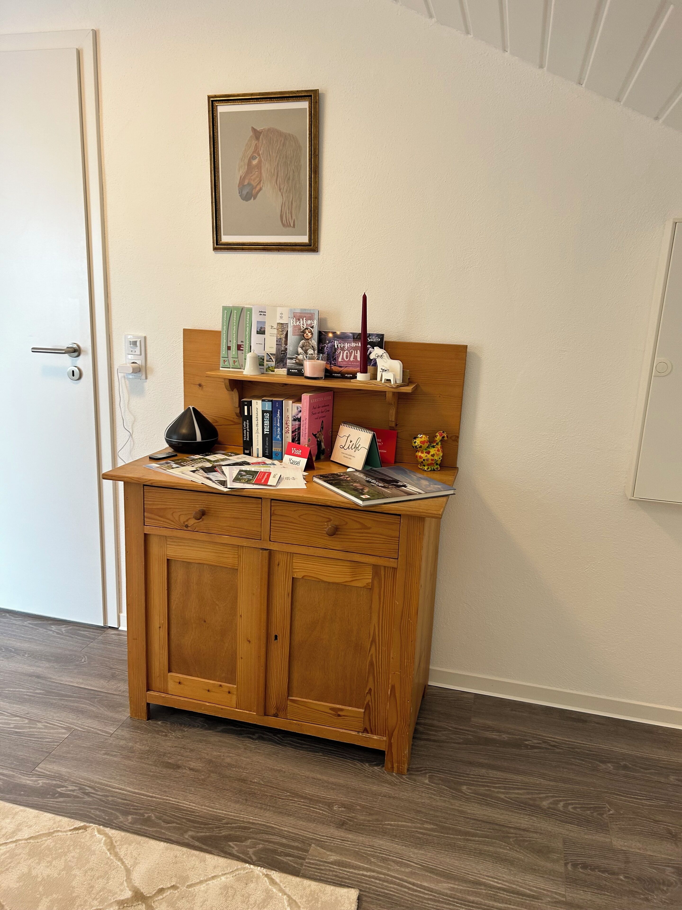 A wooden desk with a shelf above it, holding books, a candle, and a small figurine.