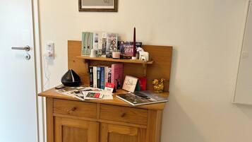 A wooden desk with a shelf above it, holding books, a candle, and a small figurine.