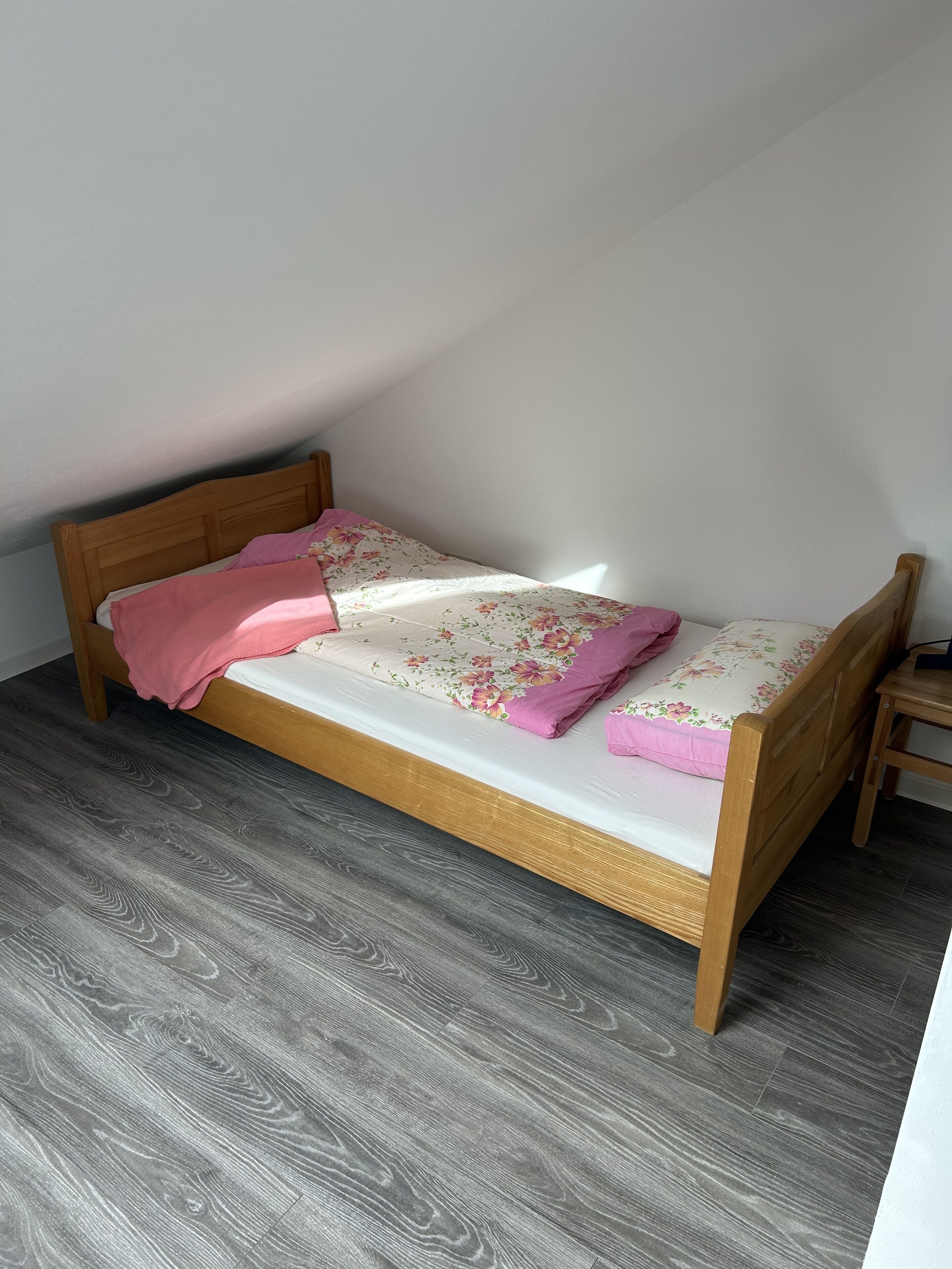 Una habitación pequeña con cama individual, estructura de madera, suelo estampado en gris y techo inclinado.