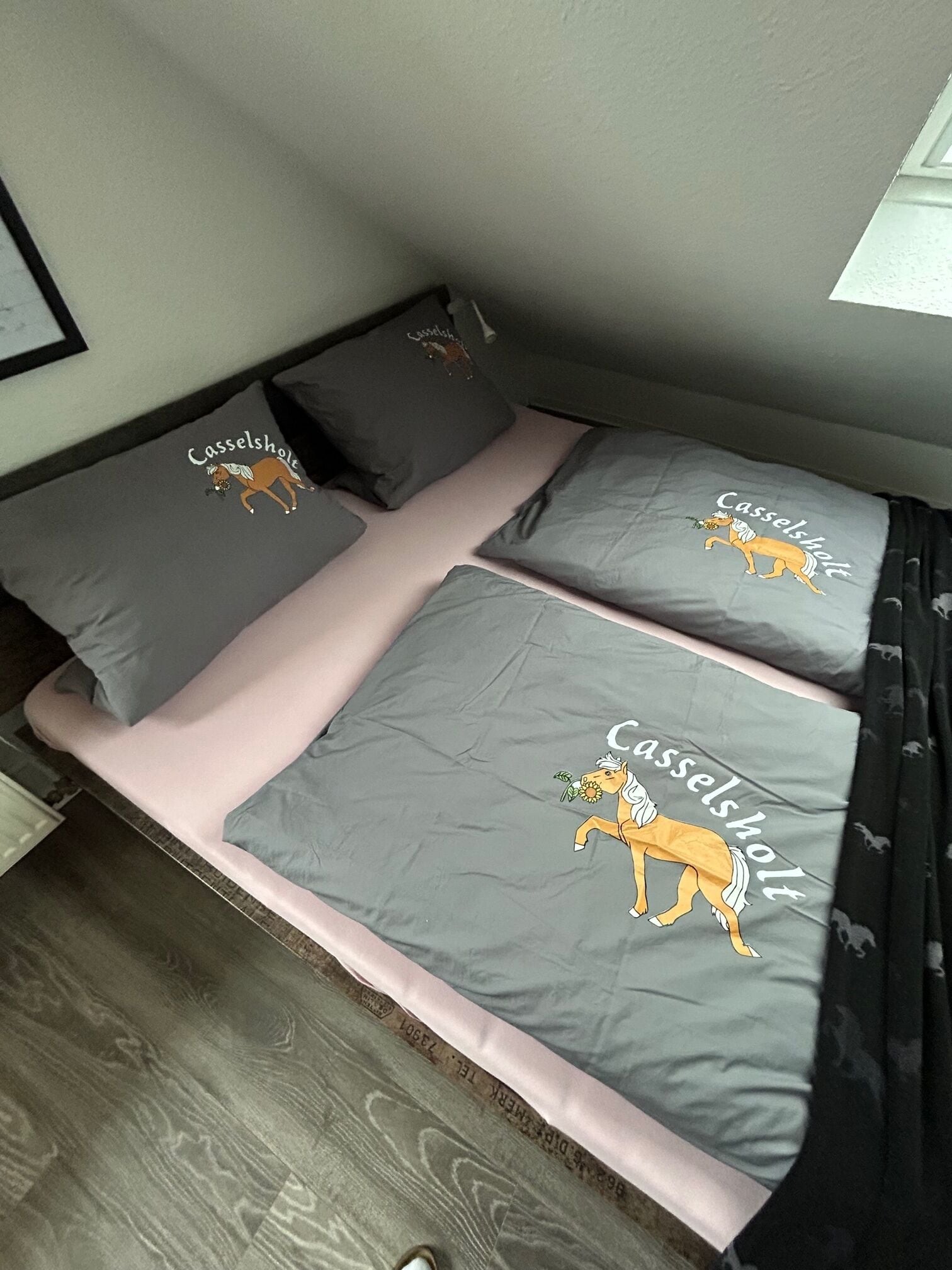 A small, cozy room with a bed featuring grey bedding with a horse illustration and the word "Casselsholt" printed on it.