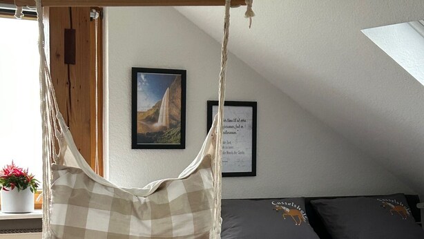 Une pièce chaleureuse avec un fauteuil en forme de hamac, un lit recouvert d’une courtepointe à motifs gris et noirs, et un tableau encadré accroché au mur.