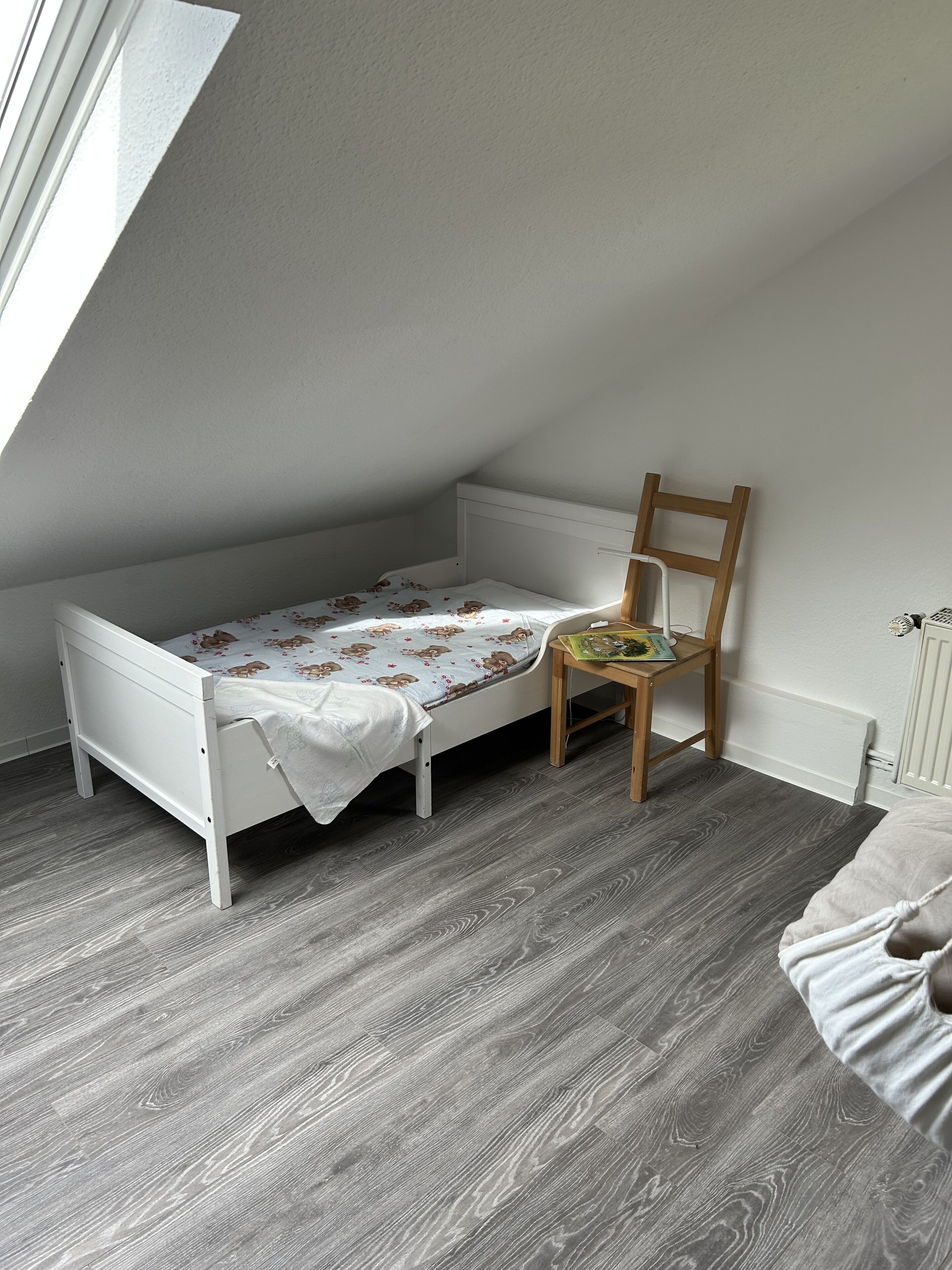 Ein kleines Zimmer mit einem Einzelbett, geneigter Decke, einem Holzstuhl und einer Heizung.