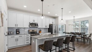 Une cuisine moderne avec des meubles blancs, un îlot central doté de tabourets de bar, des appareils en acier inoxydable et un éclairage suspendu.
