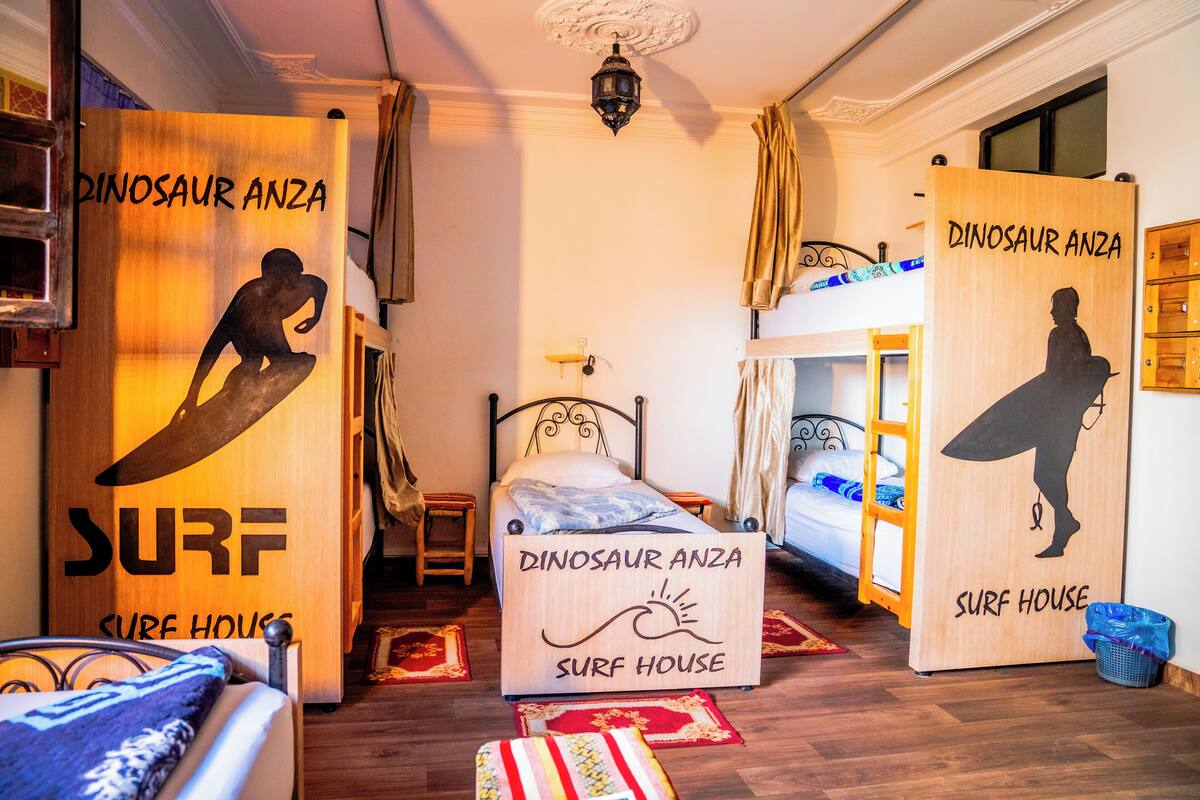 A room with bunk beds, a bed with a surf-themed headboard, and a wall with 'Dinosaur Anza Surf House' decorations.