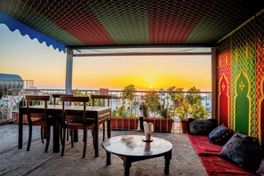 A terrace with a table, chairs, and a sofa, overlooking a cityscape at sunset. - Dinosaur anza surf house (Agadir)