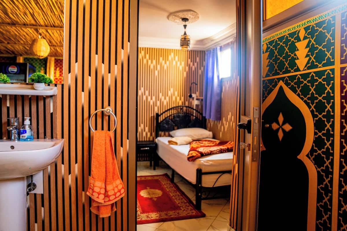 A hotel room with a bed, a bedside table, a wall-mounted shelf with toiletries, and a decorative door.