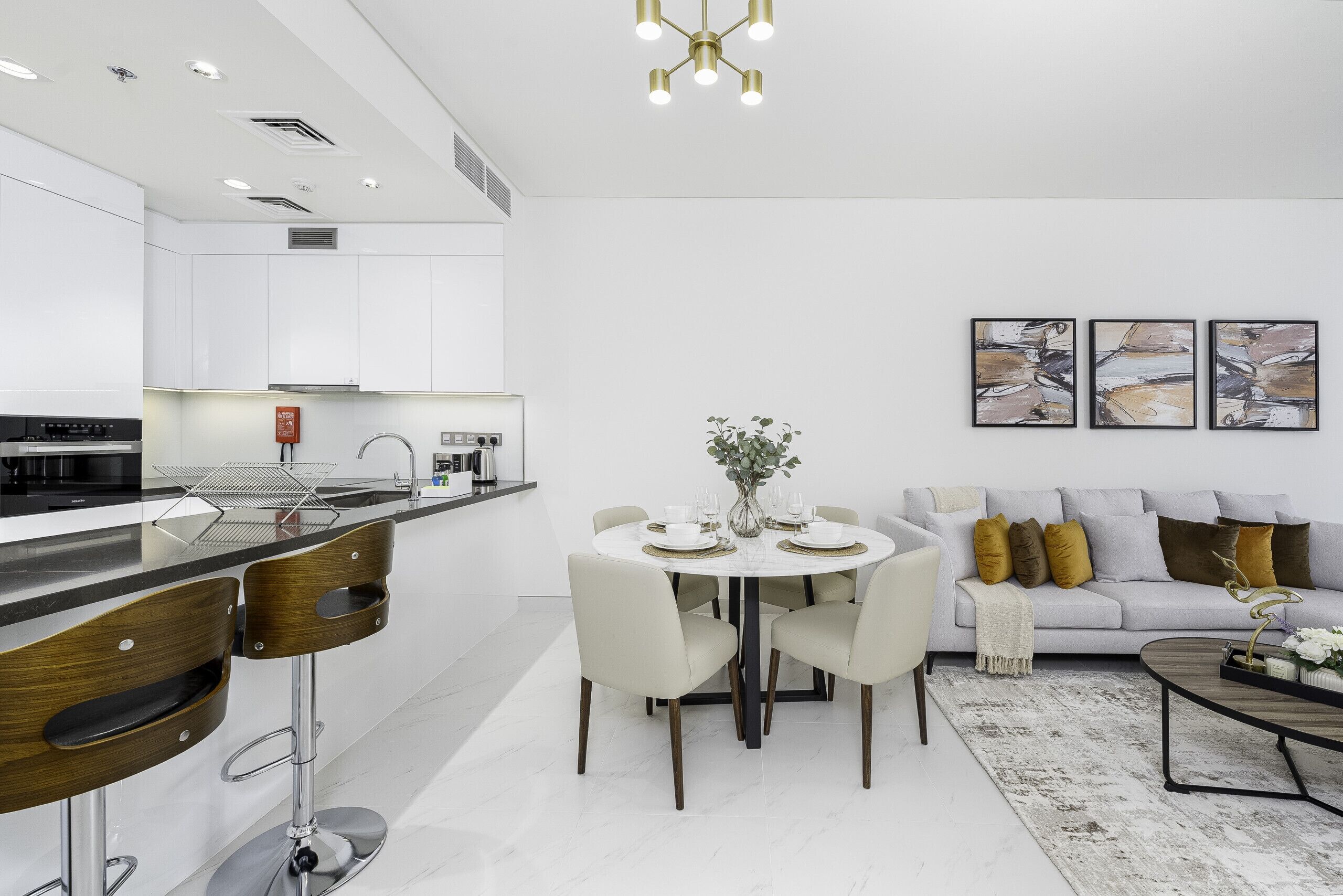 A modern kitchen with white cabinets, a dining area with a round table and chairs, a sofa with cushions, and abstract art on the wall.