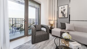 A modern living room with a grey sofa, a grey armchair, a glass door leading to a balcony, a framed abstract artwork, and a small table with a vase of flowers and a tissue box.