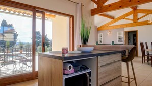 A kitchen with a large island, wooden beams, a view through the sliding glass door, and a dining area with chairs. - Villa in Provence with pool, garden and Ventoux view – Colonzelle (Colonzelle)