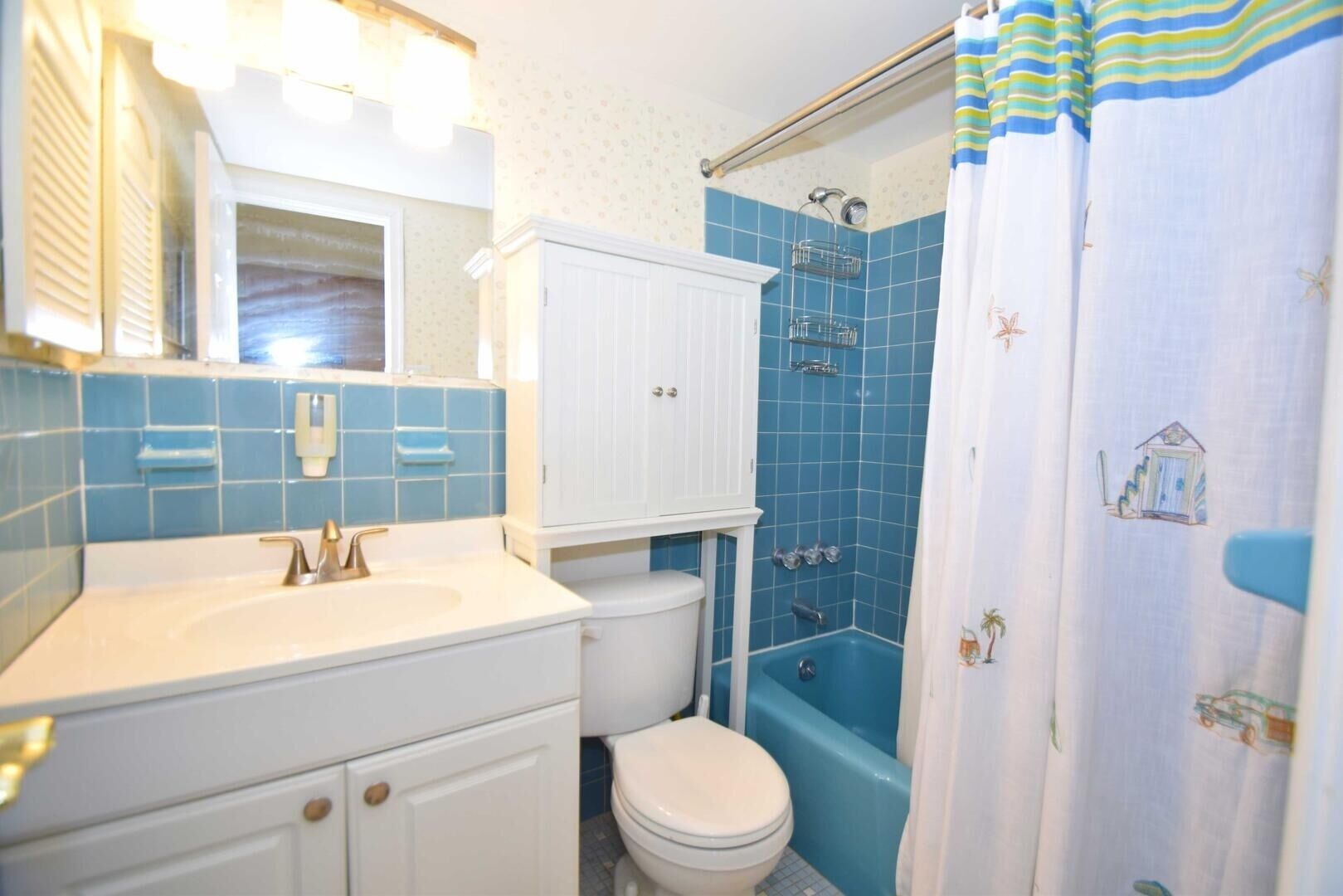 Une salle de bain avec un lavabo, des toilettes et une baignoire, tous de couleur blanche. Il y a un rideau de douche orné d'un motif représentant une plage.