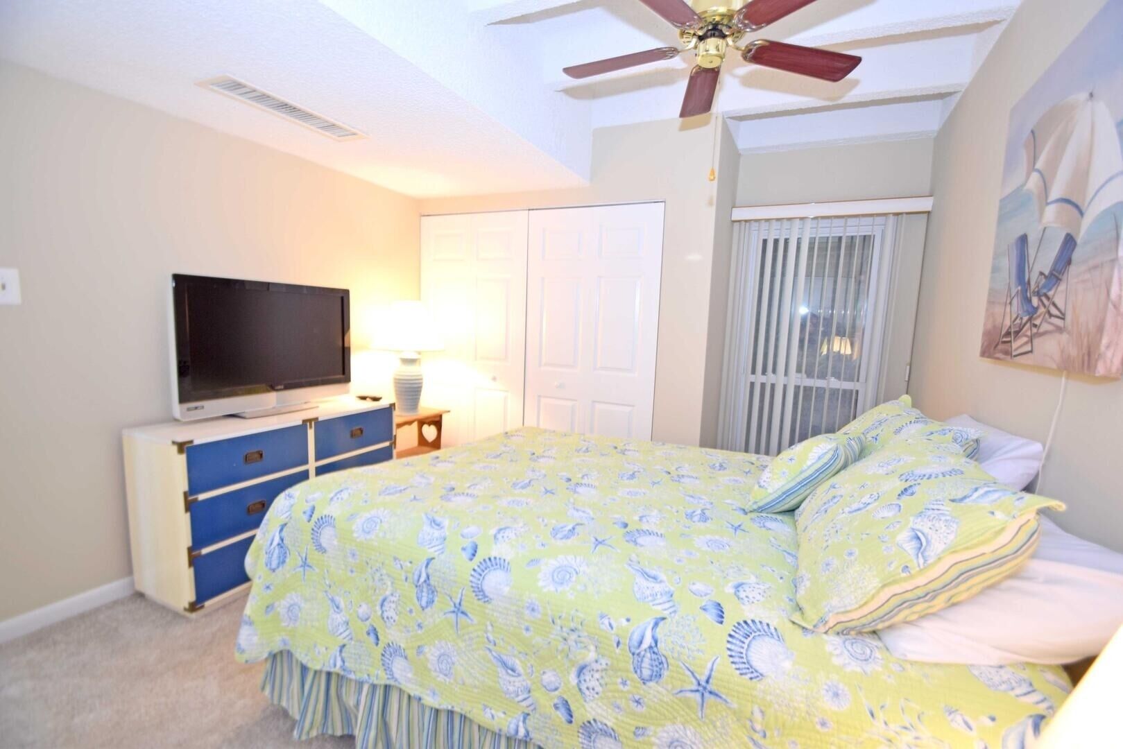 A bedroom with a bed, a television, a ceiling fan, and a window with blinds.
