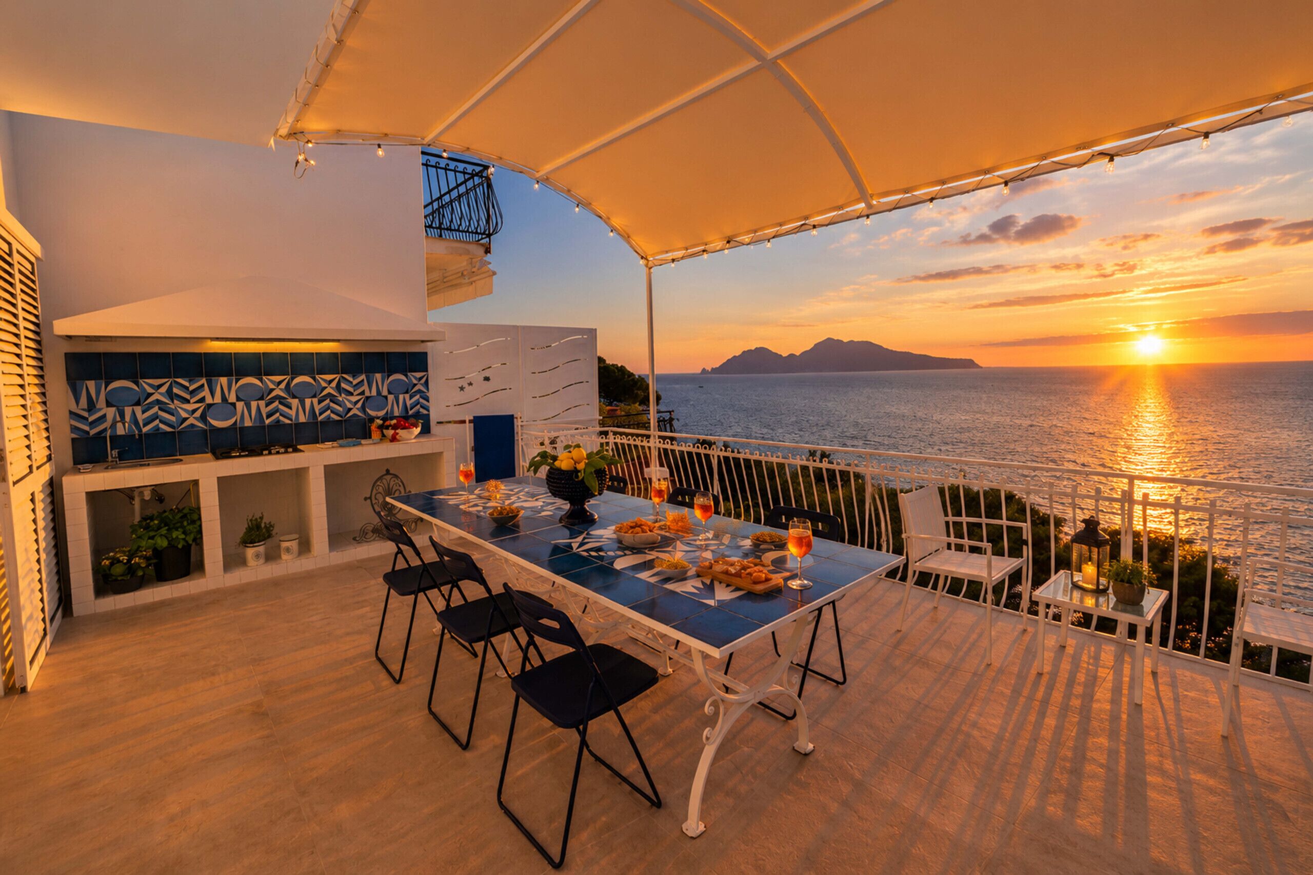 An outdoor dining area with a view of the sunset over the sea, a table set with plates and glasses, and a bar area with a tiled backsplash.
