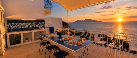 An outdoor dining area with a view of the sunset over the sea, a table set with plates and glasses, and a bar area with a tiled backsplash.
