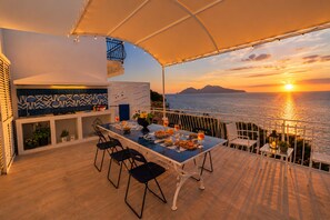 An outdoor dining area with a view of the sunset over the sea, a table set with plates and glasses, and a bar area with a tiled backsplash. - AMORE RENTALS - Villa Domus Franca with Seafront Shared Pool (Massa Lubrense)