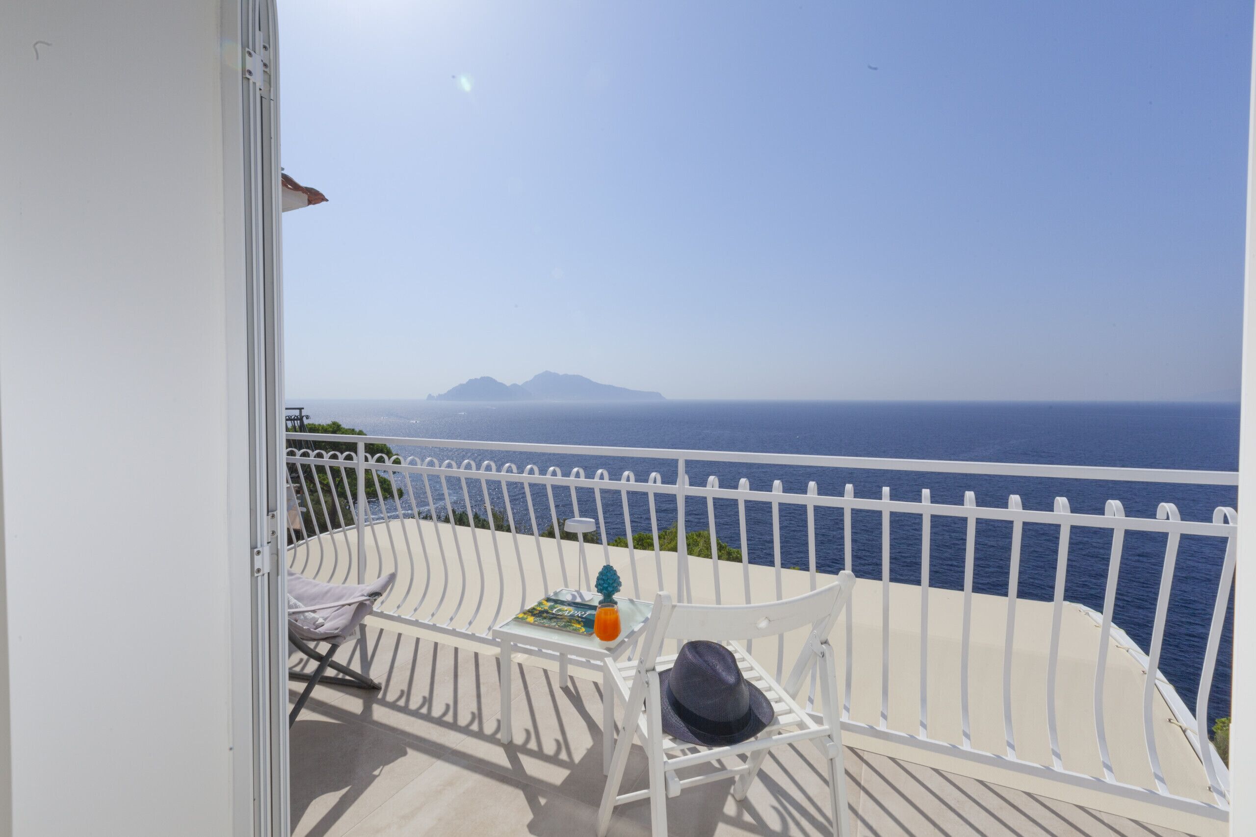 Un balcon avec une balustrade blanche, une table avec un magazine, un chapeau et une bouteille de jus d’orange.