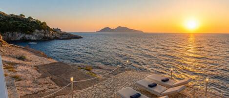 A sunset view from a terrace with lounge chairs and a rocky coastline.