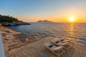 A sunset view from a terrace with lounge chairs and a rocky coastline.
