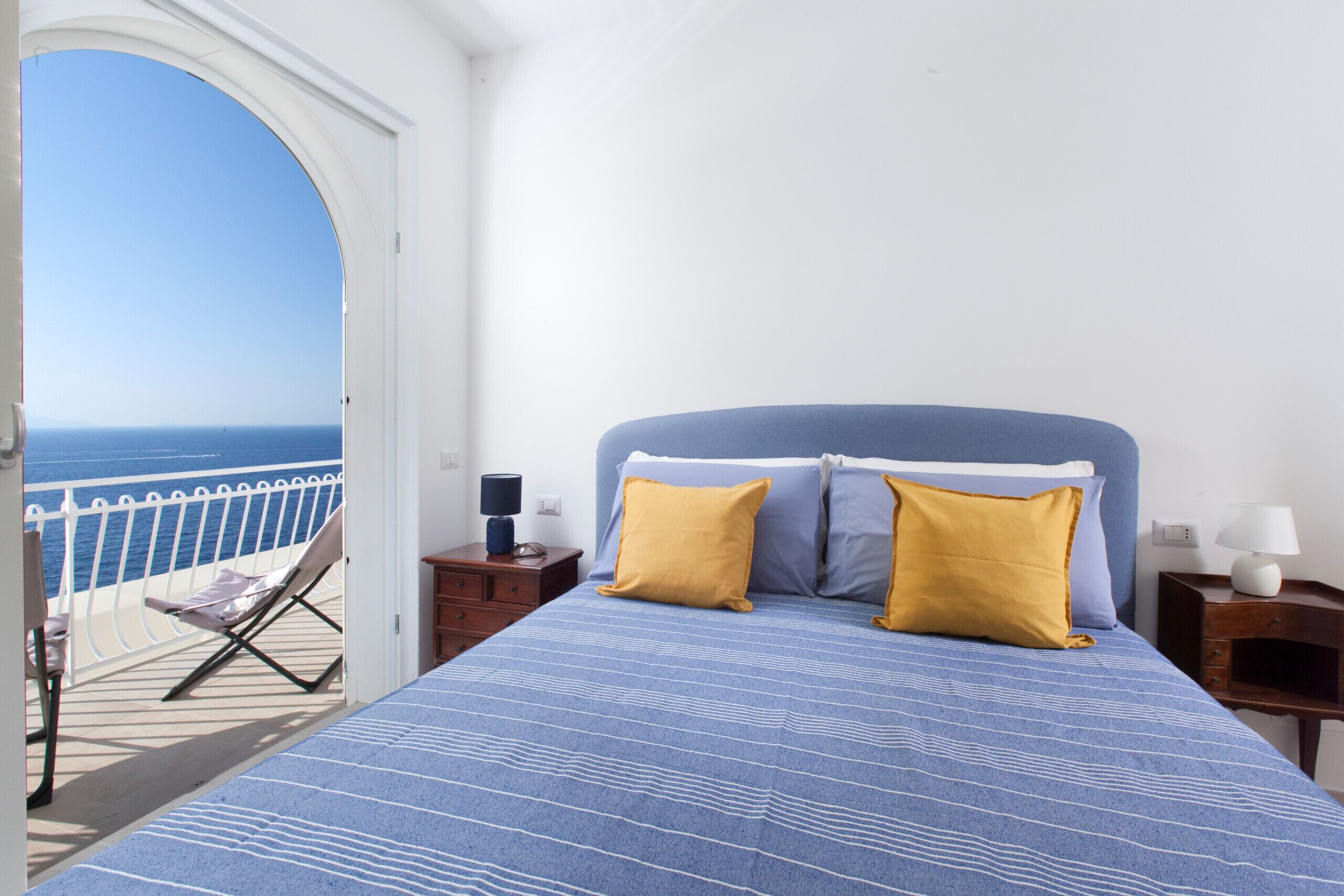 A bedroom with a bed, two pillows, a nightstand, a lamp, and a view of the sea through an open door.