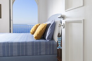 Een bed met een blauw-witte streepdeken, twee kussens (een geel en een blauw), en een sleutelhanger in de vorm van een vis op het nachtkastje.
