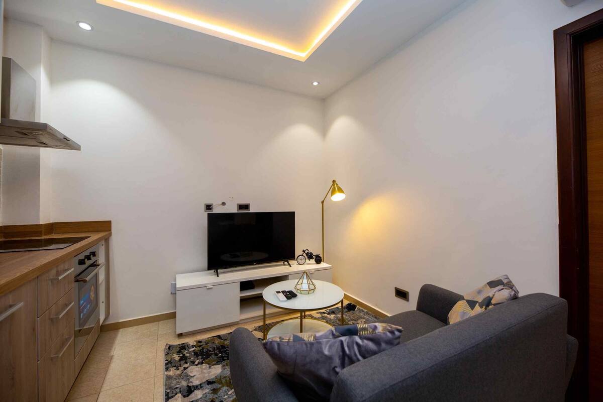 A modern living room with a sofa, a TV, a small table, and a kitchen area with cabinets and a stove.