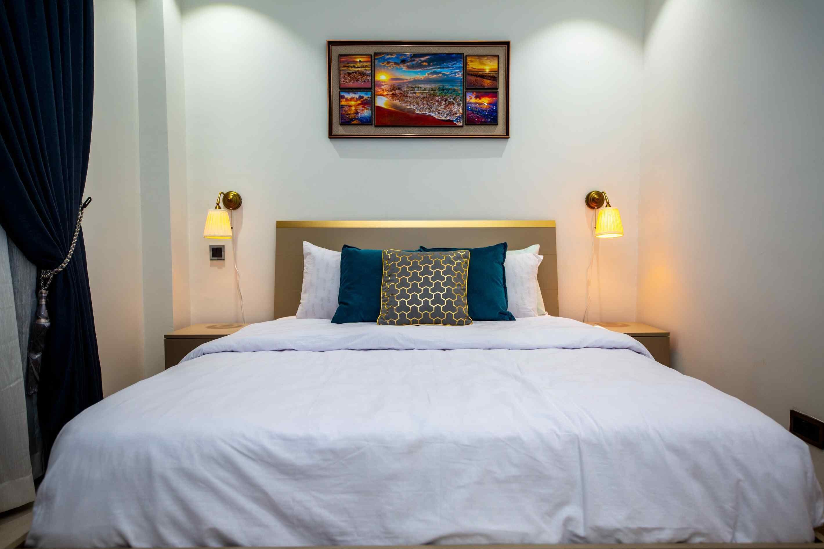 A neatly made bed with white bedding and decorative pillows, two bedside lamps, and a framed cityscape artwork above the bed.