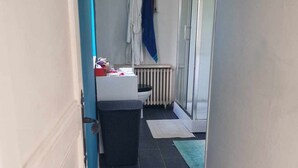 Um corredor estreito com uma porta branca à esquerda e uma porta de vidro à direita. Há um radiador na parede e um tapete azul no chão.