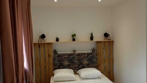 Un dormitorio con una cama, cabecera estampada con motivos florales, estante con adornos y ventana con cortinas.
