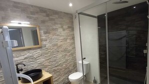 Un baño moderno con pared de piedra, inodoro, ducha con mampara de vidrio y lavabo con espejo.