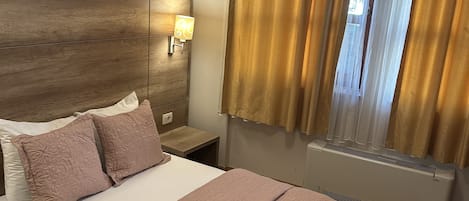 A bedroom with a wooden headboard, a bed with a patterned bedspread, a nightstand with a lamp, and a window with curtains.