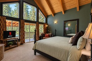 A bedroom with a large bed, a TV, a mirror, and a view of trees through the windows. - Riverside Cabin-River, Hot Tub, WIFI, Dogs OK (Leavenworth)