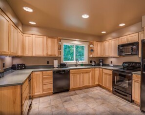 A modern kitchen with wooden cabinets, a dishwasher, a microwave, a stove, and a window with a view of greenery.